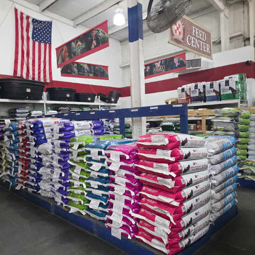 Stacks of colorful pet food bags are neatly arranged on blue shelves in a feed store. An American flag and 