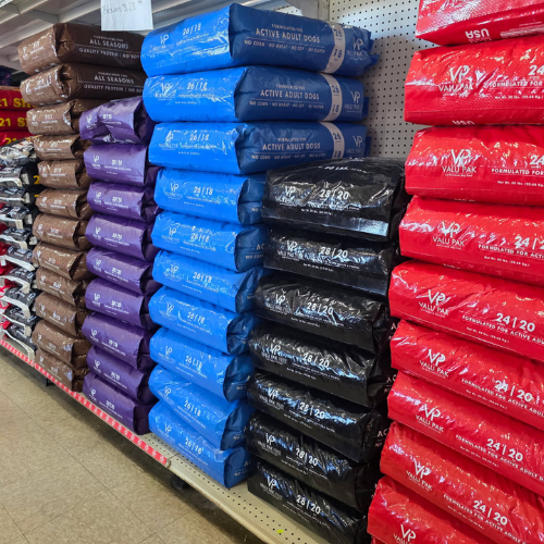 Stacks of dog food bags in a store, arranged by color: brown, purple, blue, black, and red. The display looks organized and orderly.