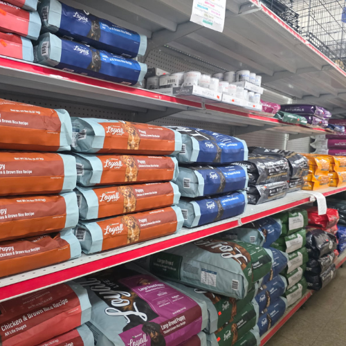 Shelves stocked with various colored bags of dog food in a store aisle. The scene conveys abundance and options, with neatly organized products.