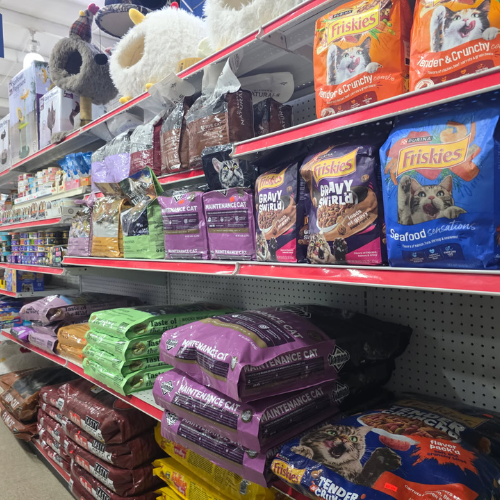 A pet store aisle filled with various colorful bags of cat and dog food, including brands like Friskies, on neatly organized red-shelved displays.