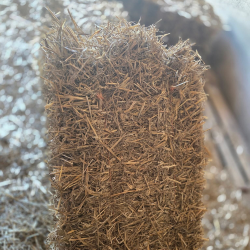 Close-up of a rectangular hay bale, showcasing tightly packed, golden-brown straw against a blurred, sunlit background, conveying rustic warmth.