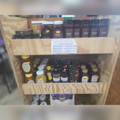 Wooden shelves displaying various honey products in jars and bottles, including clover honey and honey with pollen. Brightly lit, rustic setting.